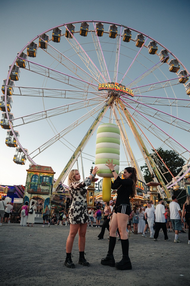 Zwei junge Frauen halten eine aufblasbare Produktnachbildung eines Twister-Eis vor einem Riesenrad auf einem Festival.